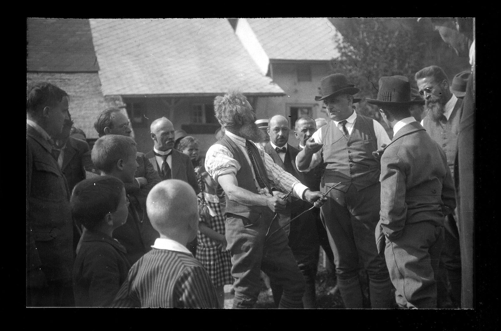 Un sourcier tient sa baguette devant une ferme, entouré d'habitants du village venus observer, en 1927